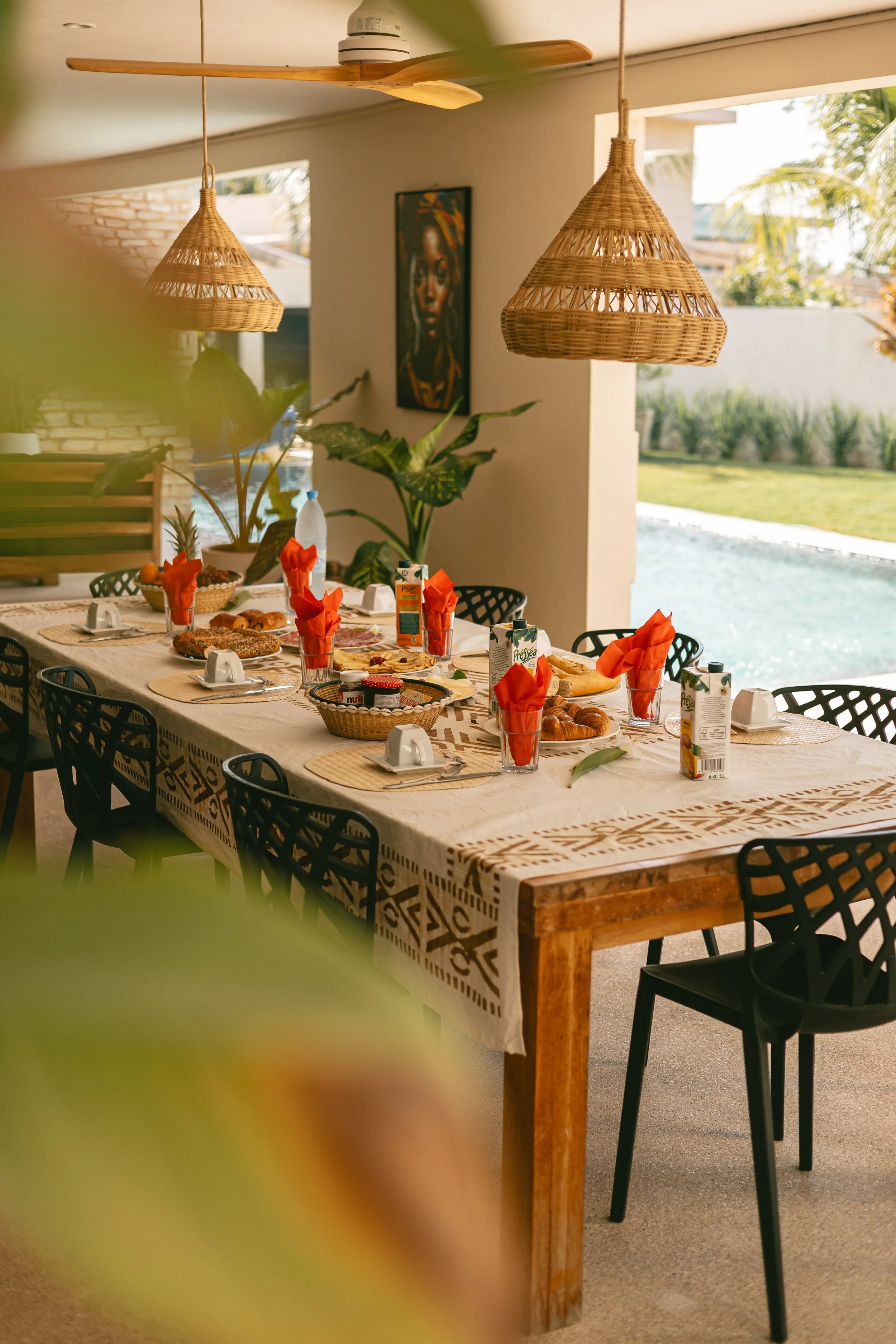 Grande table dressée avec serviettes orange et lanternes en osier, ouverte sur le jardin et la piscine