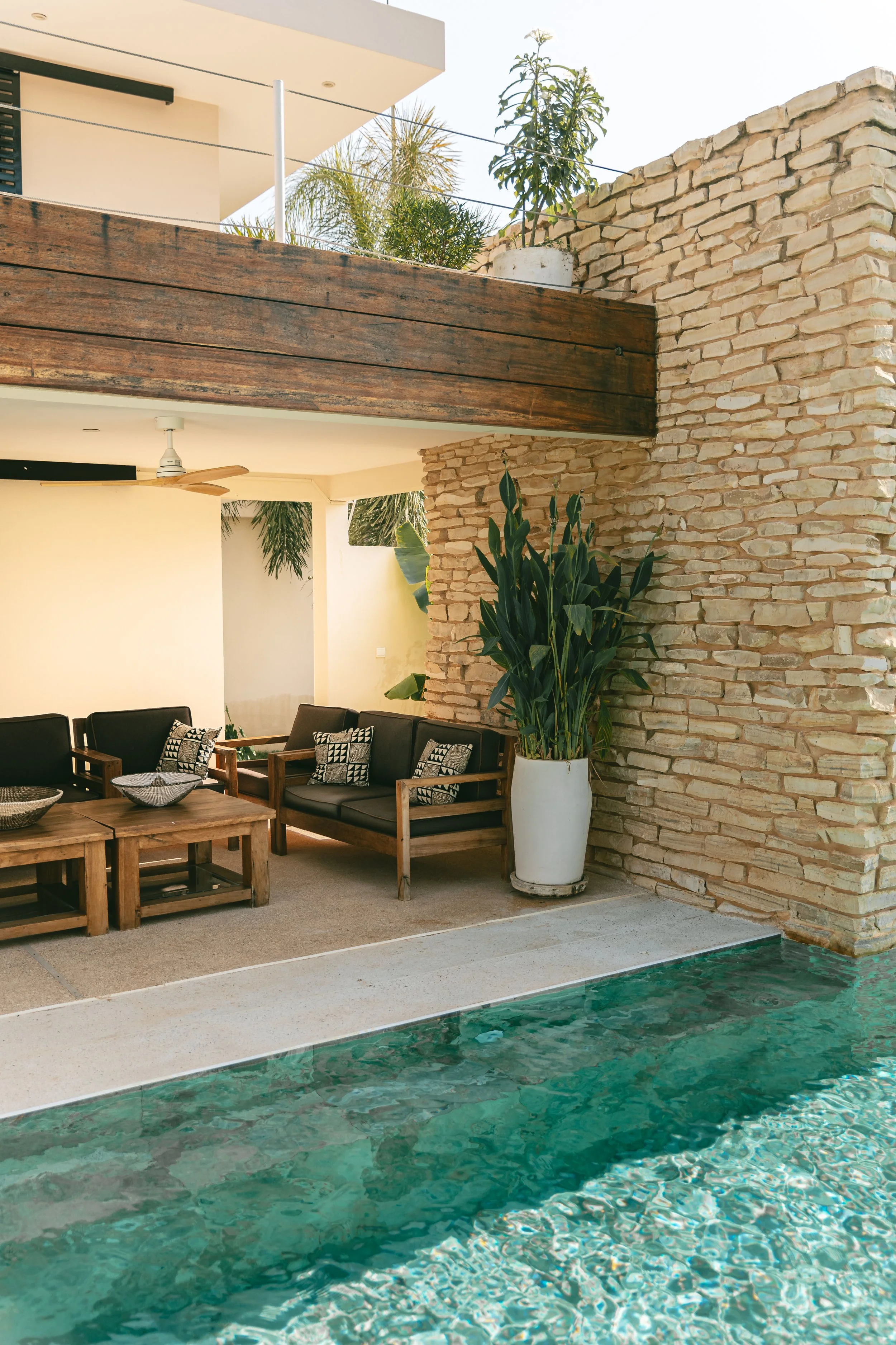 Salon extérieur sous la terrasse en bois, mur en pierre, piscine au premier plan